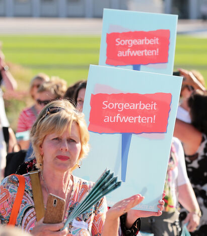 Eine Gruppe von ca. 10 Personen halten Plakate in den Händen mit den Aufschrift 'Sorgearbeit aufwerten'.