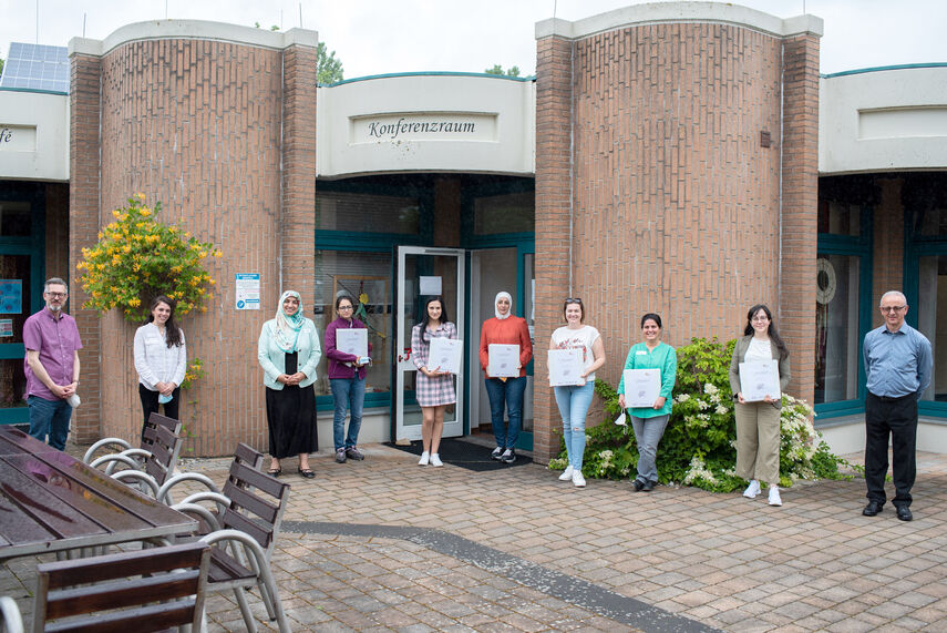 10 Personen stehen vor einem Gebäude mit der Aufschrift 'Konferenzraum'. 6 Personen halten Urkunden in der Hand.