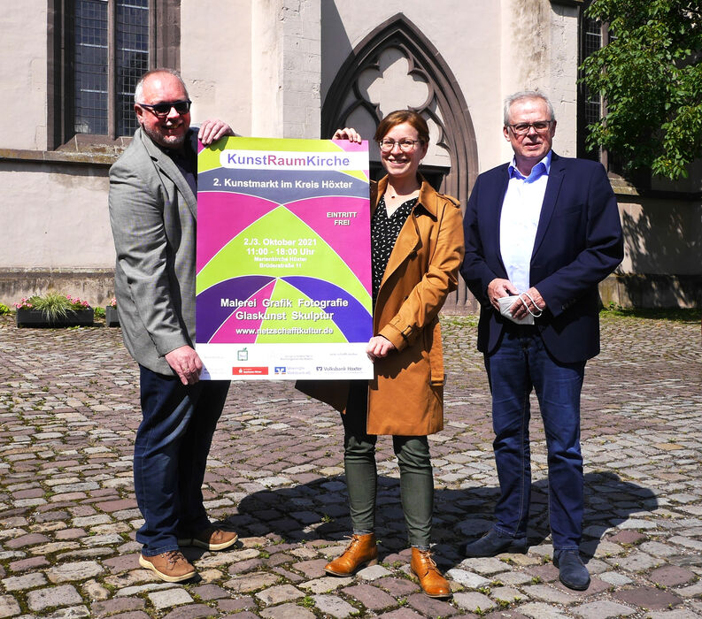 Pfarrer Uwe Neumann von der evangelischen Weser-Nethe-Kirchengemeinde, Kulturmanagerin Stephanie Koch und Kreisdirektor Klaus Schumacher präsentieren das Plakat für den 2. Kunstmarkt.