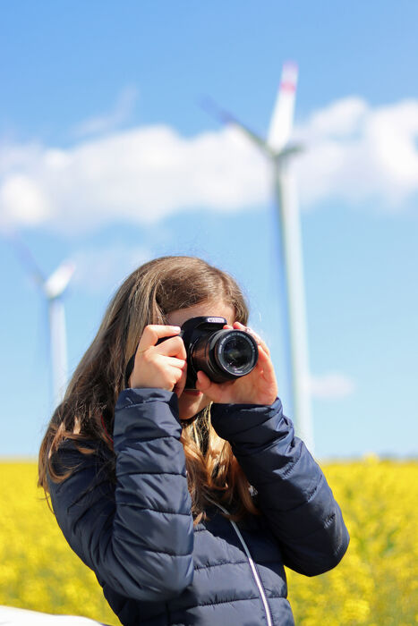 Eine junge Dame fotografiert eine Windkraftanlage. Sie steht in einem Rapsfeld.