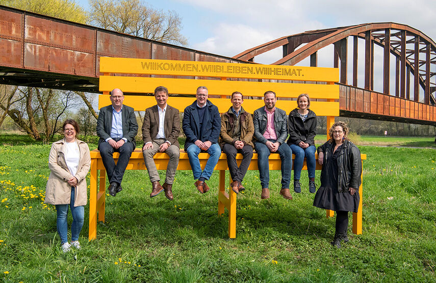 8 Personen sitzen auf einer großen Bank für der alten Eisenbahnbrücke in Corvey